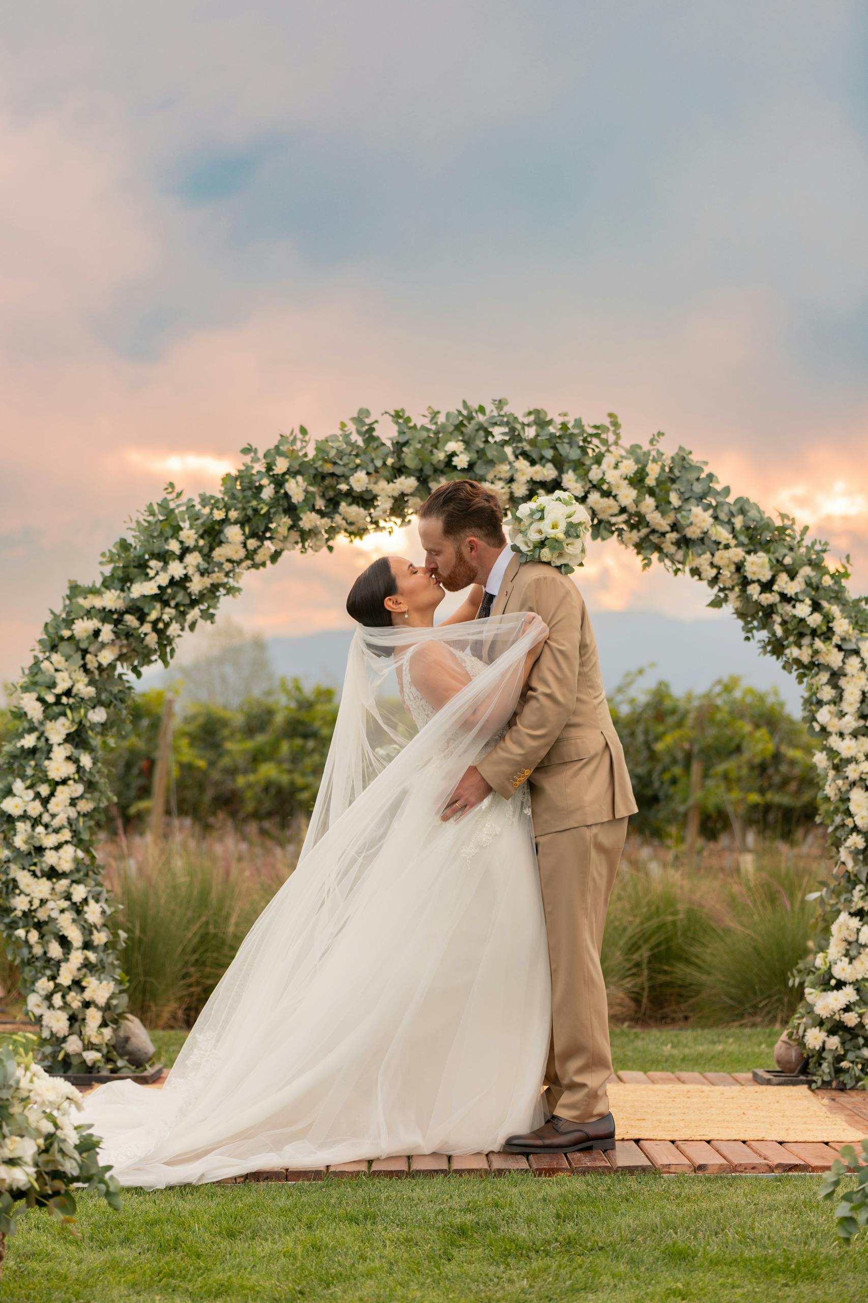 Bride and groom share a romantic kiss under floral arch in Mendoza vineyard.